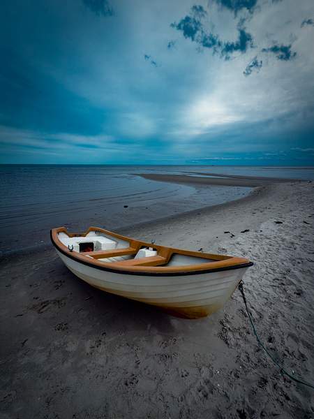 A small wooden rowboat rests on a quiet beach under a moody blue sky. The calm sea and soft light create a peaceful, almost cinematic atmosphere.
